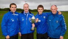 Four men in blue sports jackets stand on grass holding a trophy.