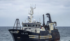 A deep-sea fishing vessel with the identification number LK 44, fitted with various antennas and equipment, sails on a calm sea under an overcast sky.