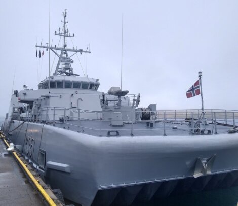 A gray naval ship with the number "605" on its side is docked. A Norwegian flag is visible at the rear of the ship. The weather is overcast.