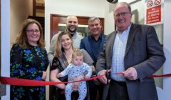 A group of six people, including a baby, is gathered inside a building. A man in a suit is cutting a red ribbon while others smile. A "No Smoking or Vaping" sign is visible in the background.