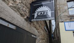 Sign for PeerieShop Café, labeled "The One Stop Coffee Shop," mounted on a stone building along a narrow alley.