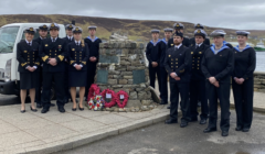 A group of naval personnel in uniform stand around a stone monument adorned with memorial wreaths, against a backdrop of hilly landscape and cloudy skies.