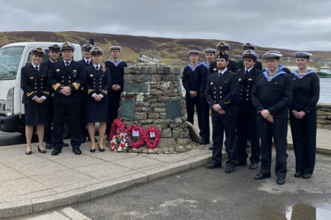 A group of naval personnel in uniform stand around a stone monument adorned with memorial wreaths, against a backdrop of hilly landscape and cloudy skies.