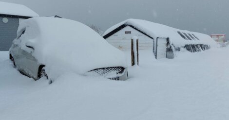 A car, a shed, and another building are covered in a thick blanket of snow, almost obscuring their shapes. The snow appears to be fresh and deep, indicating a recent heavy snowfall.