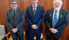 Three men in suits stand in a row in a wood-paneled room. The man on the left has gray hair and glasses, the man in the middle has dark hair and is smiling, and the man on the right has white hair and a beard.