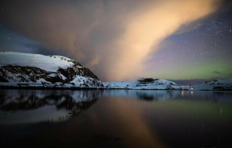 Snow-covered mountains reflecting on a calm lake under a cloudy night sky with visible stars.