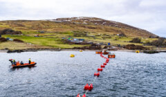 A shoreline with people working on a small boat and near red buoys in the water; a hilly landscape with scattered small structures is in the background.