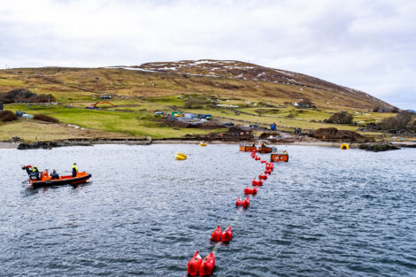 A shoreline with people working on a small boat and near red buoys in the water; a hilly landscape with scattered small structures is in the background.