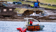 A boat, equipped with equipment and personnel in red jackets, is stationed in a body of water near a construction site with workers, machinery, and a hillside.