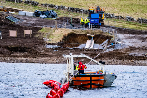 A boat, equipped with equipment and personnel in red jackets, is stationed in a body of water near a construction site with workers, machinery, and a hillside.