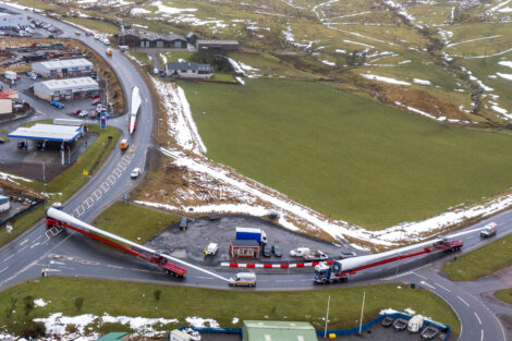 Aerial view of a large truck transporting an oversized wind turbine blade through a roundabout in a rural area with patches of snow on the ground.