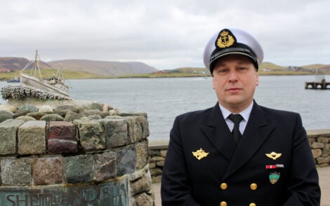 A uniformed person stands beside a stone monument with a model ship and "Shetland Bus" inscribed on it, with water and hills visible in the background.