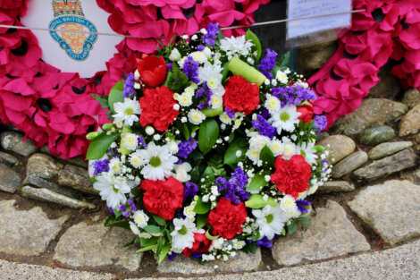 A wreath composed of red, white, and purple flowers, including carnations and daisies, is laid on a stone surface in front of a memorial adorned with pink flower decorations and a crest.