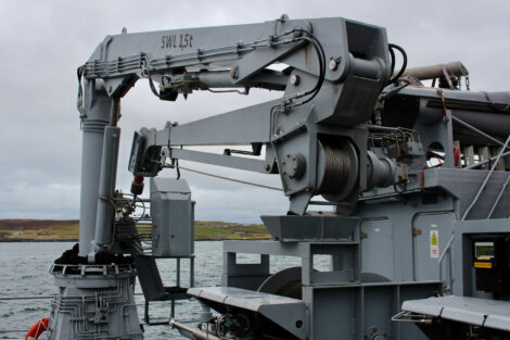 Close-up of a heavy-duty crane labeled "SWL 1.5t" mounted on a ship, with a winch and cables, set against a cloudy sky and a distant shoreline.