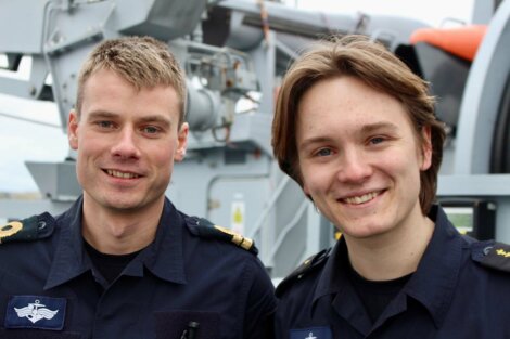 Two men in navy uniforms stand outdoors on a vessel, smiling at the camera. They are likely members of a maritime or naval service. Machinery is visible in the background.