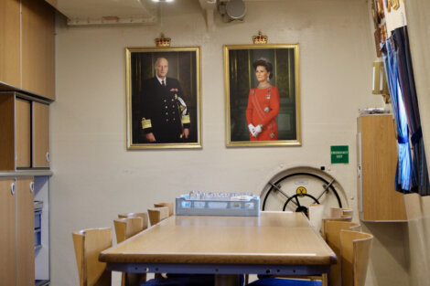 A room with a wooden table and chairs features two framed portraits on the wall. One portrait depicts a man in a military uniform, and the other shows a woman in a red dress.