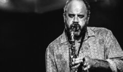 A man plays a saxophone with his eyes closed on a dark stage. He is wearing a patterned shirt and has a beard. The image is in black and white.