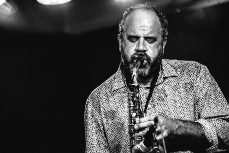 A man plays a saxophone with his eyes closed on a dark stage. He is wearing a patterned shirt and has a beard. The image is in black and white.