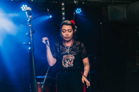 Person holding a microphone and performing on stage under blue lights, with a tiger design on their black t-shirt and a red hair accessory.