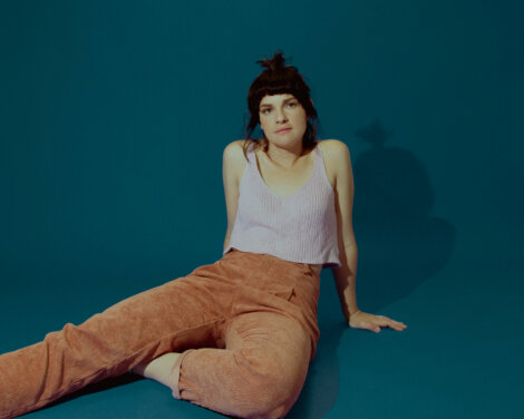 A person with short, dark hair sits on the floor against a teal background, wearing a white sleeveless top and brown pants.