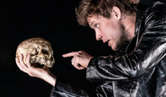 A person wearing a black jacket holds a skull and points at it with a serious expression.