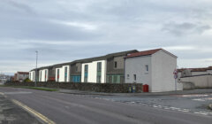 A street corner with a row of modern, two-story houses with flat and sloped roofs. The road is empty, and the sky is overcast. A traffic sign and mailbox are visible on the right.