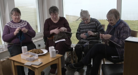 Four elderly women are sitting in a room, knitting. A table in front of them holds cups, plates, and snacks. It appears to be a casual gathering in a bright room with large windows.