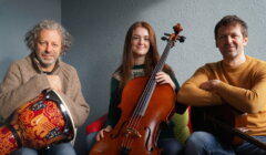 Three people seated indoors; the person on the left holds a drum, the center person holds a cello, and the person on the right holds a guitar.