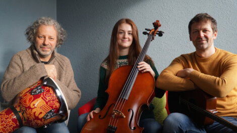 Three people seated indoors; the person on the left holds a drum, the center person holds a cello, and the person on the right holds a guitar.