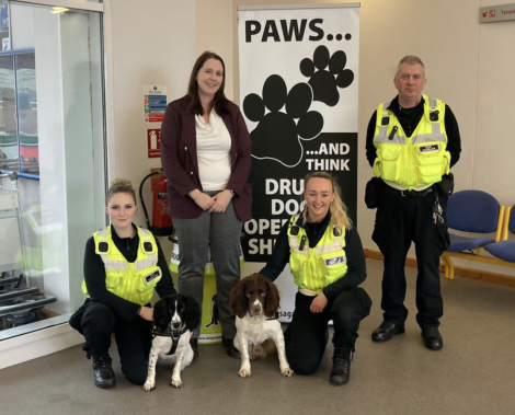 Four police officers, three wearing reflective yellow vests, pose with two dogs in front of a banner that reads "Paws...and think" and "Drug dogs operate here.