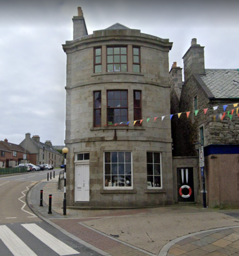 A three-story stone building with arched windows, bunting overhead, and a road curving around it. The building has a white door and a display window with decorations.