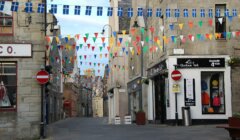 A narrow street in a small town decorated with colorful pennant flags and two no entry signs. Buildings on both sides include a store displaying outdoor gear.