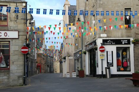 A narrow street in a small town decorated with colorful pennant flags and two no entry signs. Buildings on both sides include a store displaying outdoor gear.