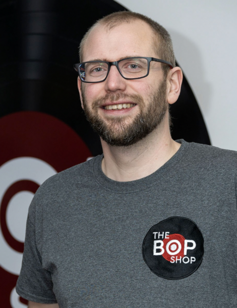 A smiling man with glasses and a beard is wearing a grey T-shirt with a logo that says "The Bop Shop" in front of a large vinyl record decoration.