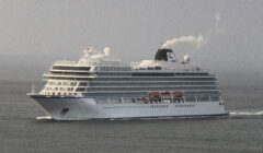 A large cruise ship with multiple decks and lifeboats sails through calm waters under a grey sky. Steam emits from its smokestack.