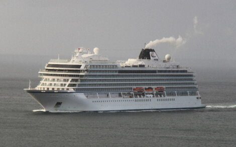 A large cruise ship with multiple decks and lifeboats sails through calm waters under a grey sky. Steam emits from its smokestack.