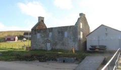 An old stone building with a partially collapsed roof stands next to a modern structure near a dirt road and grassy area by the beach. There are houses on a hill in the background.