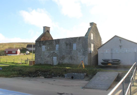 An old stone building with a partially collapsed roof stands next to a modern structure near a dirt road and grassy area by the beach. There are houses on a hill in the background.