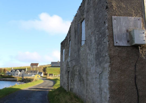 A weathered stone building with boarded-up windows stands by a narrow, paved path leading to several houses in the distance under a clear sky.