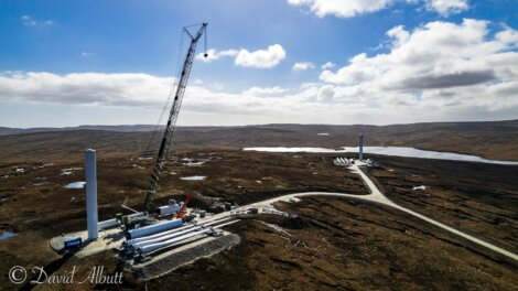 A crane and construction materials are present at a wind farm development site in a hilly and remote area, with several wind turbine components visible near a dirt road winding through the landscape.