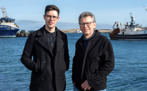 Two men in dark jackets stand by the water, with fishing boats docked in the background on a clear day.