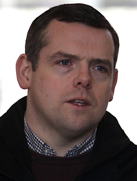 A man with short dark hair and light skin wearing a black jacket and checked shirt, looking slightly to the side with a neutral expression.