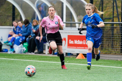 Two female soccer players chase a ball on a field, one in a pink jersey and the other in a blue jersey. Spectators and teammates are visible in the background on the sidelines.