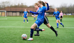 Two girls are playing soccer; one in a blue uniform is attempting to kick the ball while a girl in a pink and black uniform is closely defending. Other players and a goalpost are visible in the background.