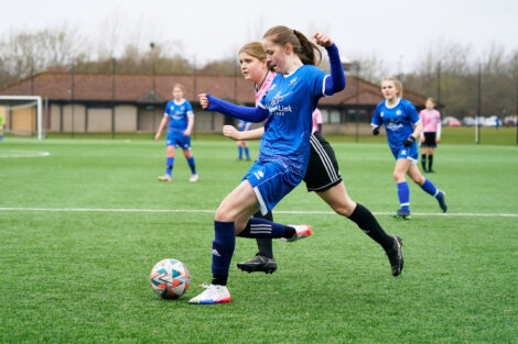 Two girls are playing soccer; one in a blue uniform is attempting to kick the ball while a girl in a pink and black uniform is closely defending. Other players and a goalpost are visible in the background.