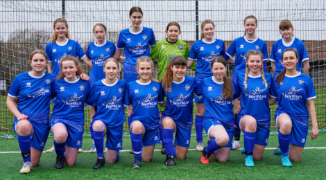 A girls' soccer team in blue uniforms poses for a group photo on a field with a goal and net in the background.