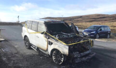 A severely burnt SUV with caution tape around it sits on a roadside with minimal traffic. Another vehicle is parked nearby, and the landscape is mostly open with hills and a cloudy sky.