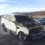 A severely burnt SUV with caution tape around it sits on a roadside with minimal traffic. Another vehicle is parked nearby, and the landscape is mostly open with hills and a cloudy sky.