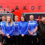 Seven individuals stand in front of a red wall with the word "Malakoff" and two vans branded "Malakoff Shetland," wearing blue and black company uniforms.