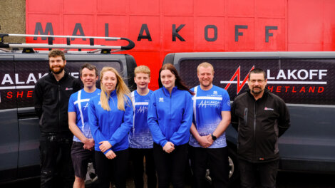 Seven individuals stand in front of a red wall with the word "Malakoff" and two vans branded "Malakoff Shetland," wearing blue and black company uniforms.